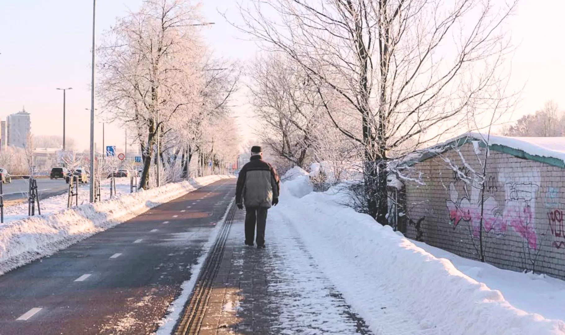 A person walking alone on a snowy, icy pathway beside a road on a winter morning, with frost-covered trees and snowbanks creating hazardous conditions that increase the risk of a fall on ice and potential personal injury.