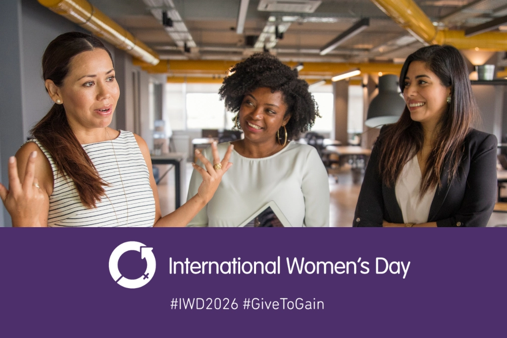 Three professional women standing together and smiling in an office setting, above a purple International Women’s Day banner featuring the IWD symbol and the words International Women’s Day with hashtags IWD2026 and GiveToGain, used by Gildeas Solicitors for International Women’s Day 2026.