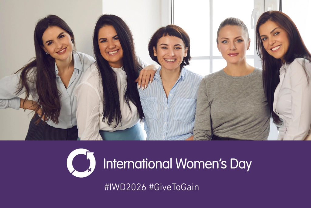 Five professional women standing together and smiling in an office setting, above a purple International Women’s Day banner featuring the IWD symbol and the words International Women’s Day with hashtags IWD2026 and GiveToGain, used by Gildeas Solicitors for International Women’s Day 2026.